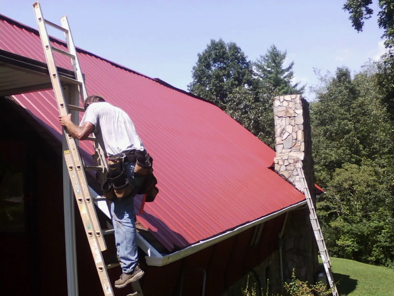 Roofer on a ladder beside a red metal roof and stone chimney — Soffit & Fascia Repair by Roofing Brothers in Stanardsville